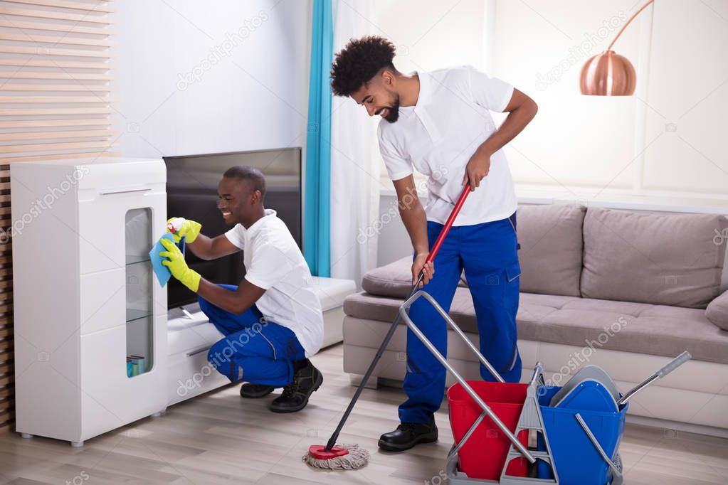 Two Smiling Young Male Janitor Cleaning The Cabinet And Mopping Floor In The Living Room