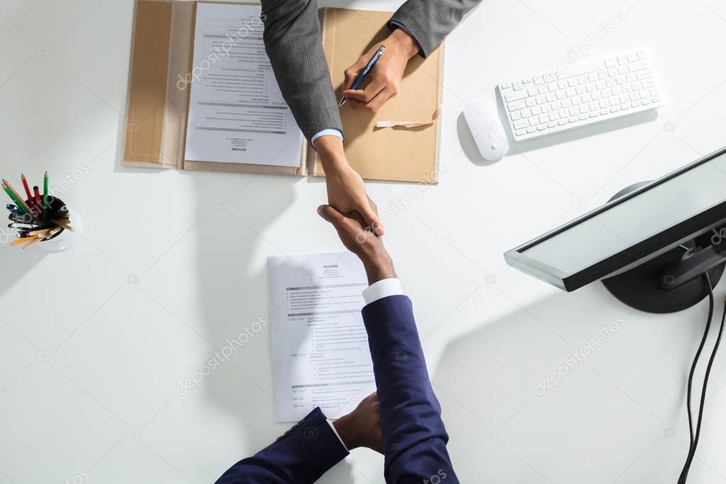 High Angle View Of Businessperson Shaking Hand With Candidate Over White Desk