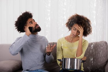 Young Man Calling Plumber While Woman Using Utensil For Collecting Water Leaking From Ceiling clipart