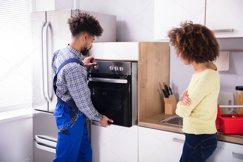 Woman Looking At Young Repairman Fixing Oven In Kitchen