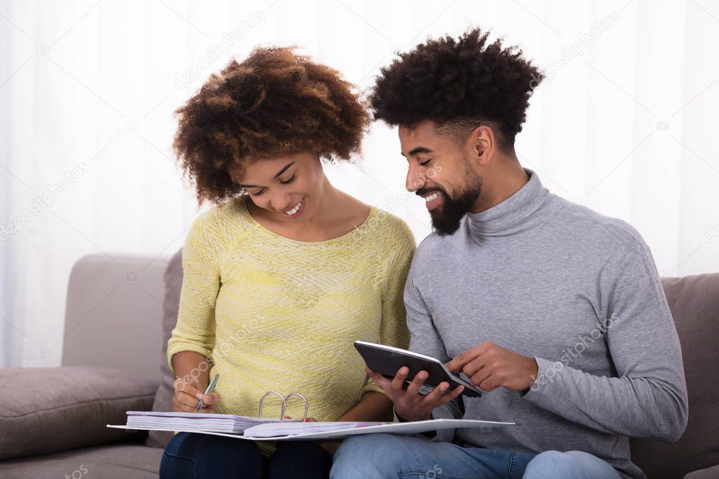 Young Couple Sitting On Sofa Calculating Bill With Calculator