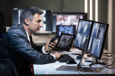 Businessman Talking On Walkie Talkie While Looking At CCTV Camera Footage On Multiple Computer Screen clipart