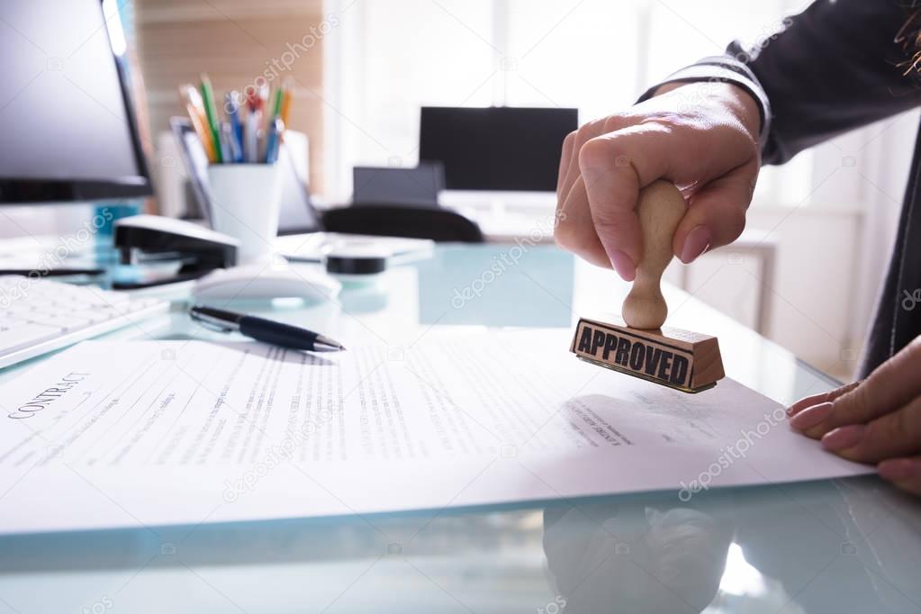 Close-up Of A Businessperson's Hand Stamping Approved On Contract Paper In Office