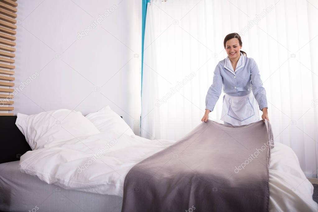 Smiling Female Housekeeper Making Bed In Hotel Room