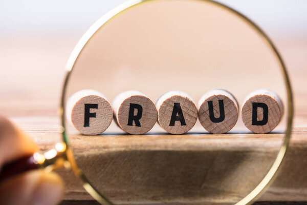 Close-up Of A Person's Hand Examining Fraud Blocks Through Magnifying Glass On Wooden Desk
