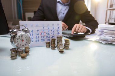 Close-up Of Stacked Coins And Calendar Besides Businessperson's Hand Using Calculator clipart