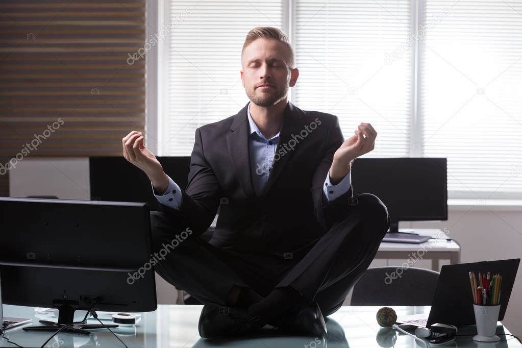 Young Businessman Sitting On Desk Meditating In Office