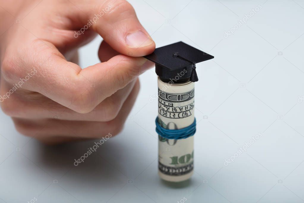 Close-up Of A Person's Hand Placing Black Graduation Hat Over American Banknotes