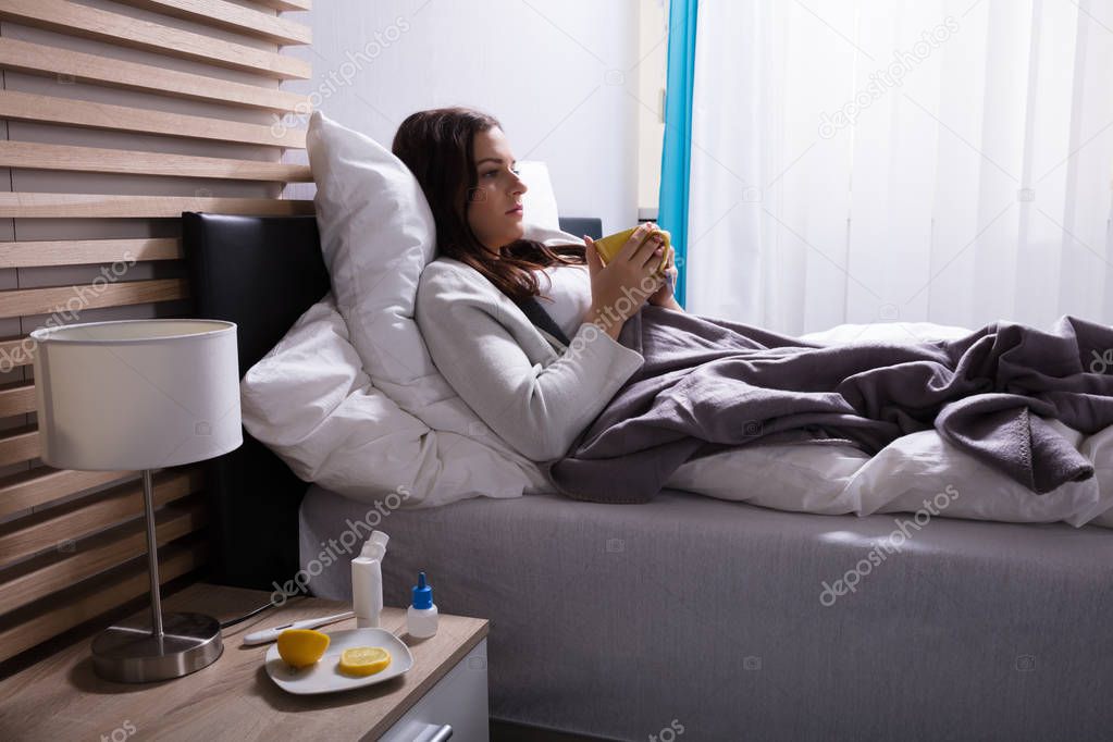 Sick Woman Lying On Bed Drinking Cup Of Tea