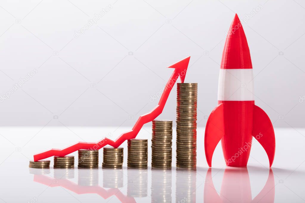 Close-up Of Red Rocket Besides Stacked Coins And Arrow Showing Upward Direction