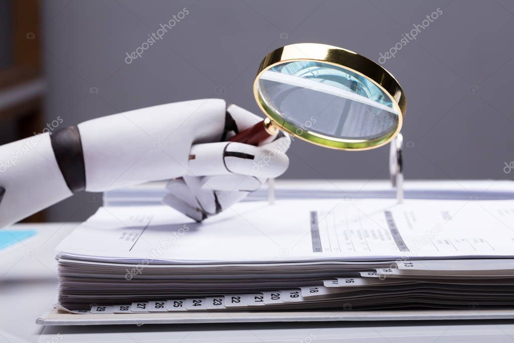 Close-up Of A Robotic Hand Holding Magnifying Glass Over Invoice At Workplace