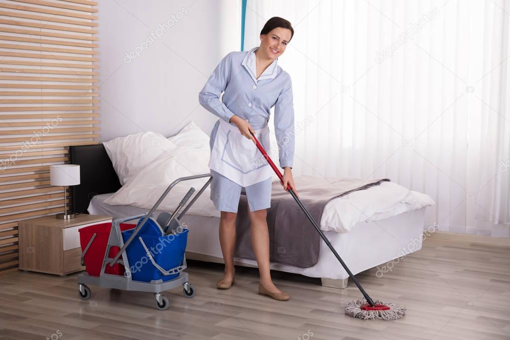 Young Female Housekeeper Cleaning Floor With Mop In Bedroom