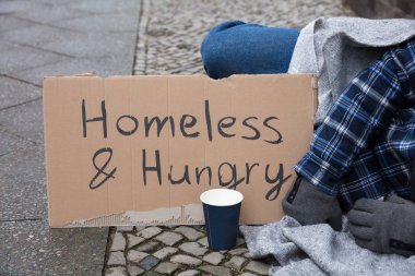 Male Beggar Lying On Street With Homeless And Hungry Text On Cardboard clipart