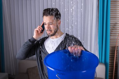 Worried Young Man Calling Plumber While Leakage Water Falling Into Bucket At Home clipart