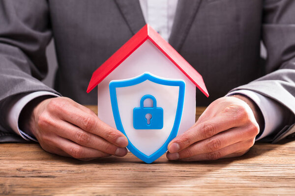 Close-up Of A Businessperson's Hand Holding Shield Security Icon Besides House Model