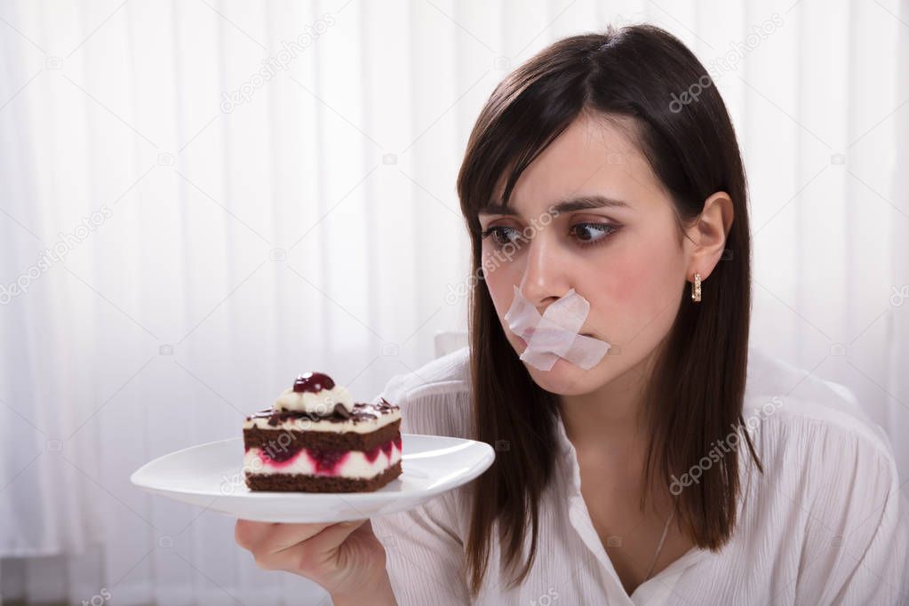 Young Woman With Sticky Tape Over Her Mouth Holding Slice Of Cake On Plate