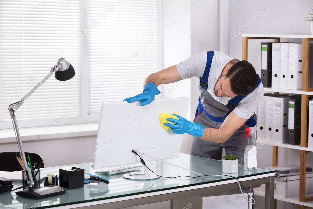 Young Male Janitor Cleaning Computer With Sponge In Office