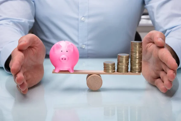 Businessperson\'s Hand Protecting Balance Between Pink Piggybank And Increasing Stacked Coins On Seesaw