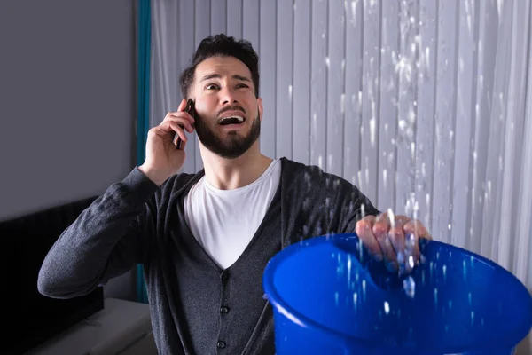 Worried Young Man Calling Plumber While Leakage Water Falling Bucket — Stock Photo, Image