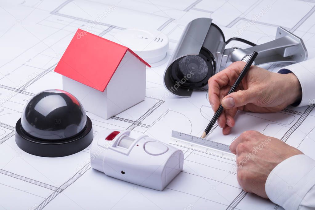 Close-up Of An Architect's Hand Drawing Blueprint Near Security Equipments And House Model