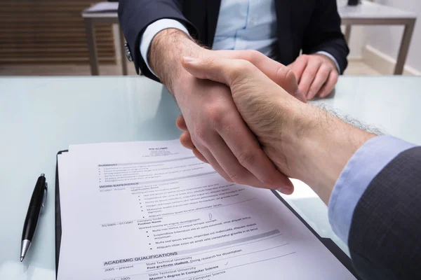 Businessperson And Applicant Shaking Hand With Resume On Desk