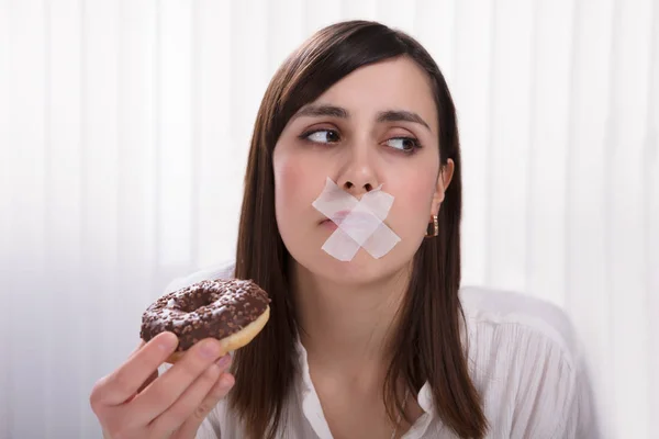 Close-up Of A Young Woman With Sticky Tape Over Her Mouth Unable To Eat Donut