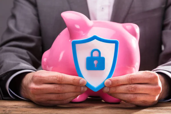 Businessperson's Hand Holding Shield Security Icon In Front Of Pink Piggybank