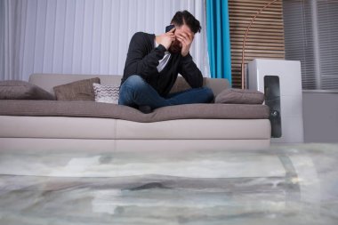 Sular altında su kaçağı için tesisatçı çağıran kanepede oturan adam önünde yerden