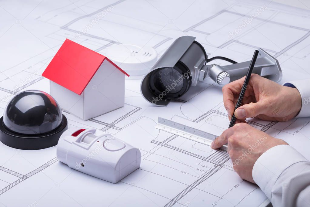 Close-up Of An Architect's Hand Drawing Blueprint Near Security Equipments And House Model