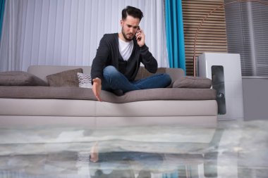 Flooded Floor From Water Leakage In Front Of Man Sitting On Sofa Calling To Plumber clipart