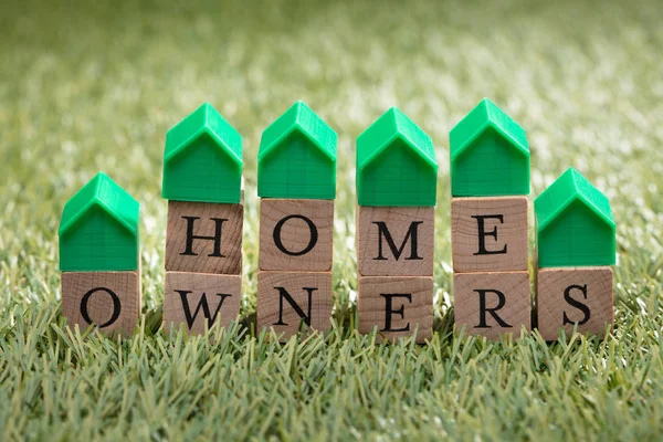 Miniature House Model Over Wooden Block Showing Home Owners Text On Green Grass