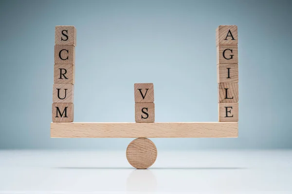 Balance Of Wooden Scrum Vs Agile Blocks On Seesaw Over The Desk Against Blue Background