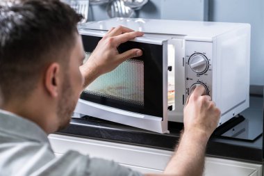 Young Man Preparing Food In Microwave Oven On Kitchen Counter clipart