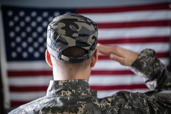 Portrait Of Serious Solider Standing In Front Of Us Flag Saluting