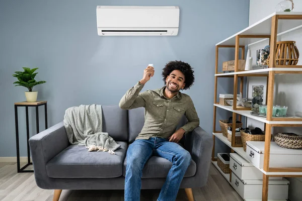 Joven Hombre Feliz Sentado Sofá Funcionamiento Aire Acondicionado Con Control —  Fotos de Stock