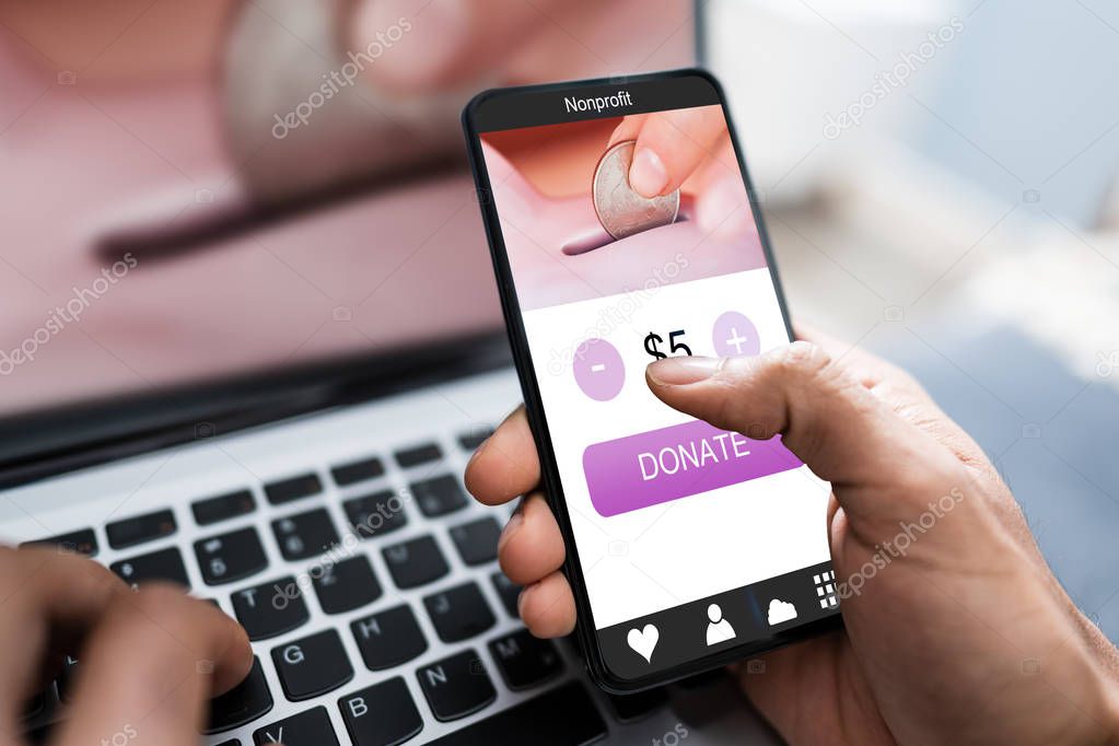 Man's Hand Donating Money On Mobile Phone Over Laptop