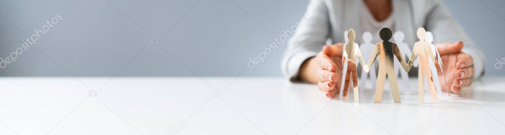 Close-up Of A Businessperson's Hand Protecting Cut-out Figures On Desk