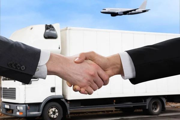 Businessmen Shaking Their Hands In Front Of A Commercial Land Vehicle And Flying Airplane Outdoors