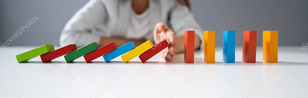 High Angle View Of A Businessperson Stopping Colorful Dominoes From Falling On Desk