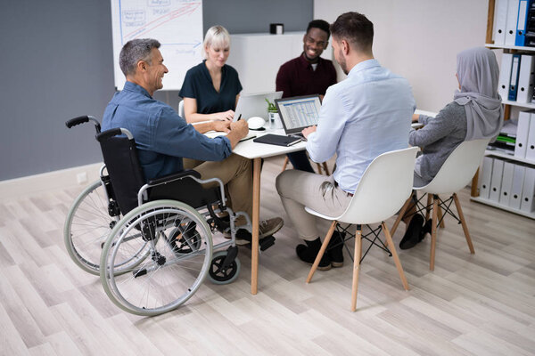 Disabled Mature Businessman Sitting At Desk With Laptop In Conference Room