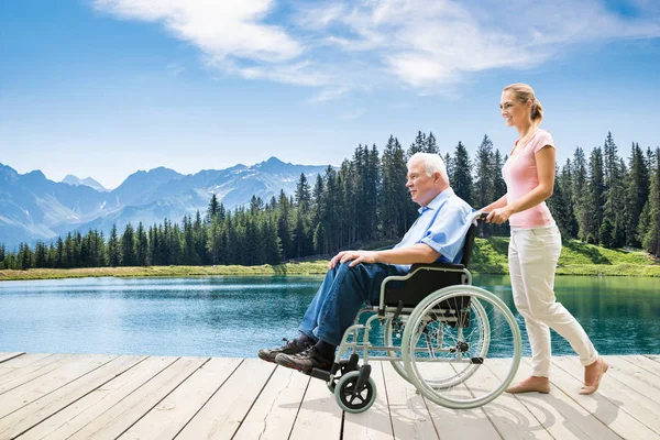 Young Woman Assisting Her Disable Grandfather In Wheelchair Walking On Boardwalk Near Lake