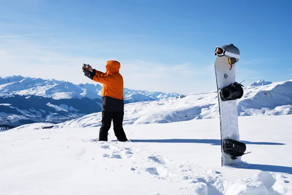 スノーボードや冬のスポーツアクセサリーの近くの雪景色を携帯電話で写真を撮る男 — ストック写真