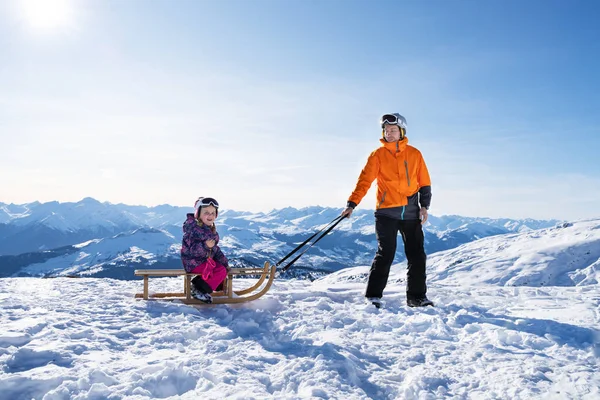 雪の中で木のそりで娘を引っ張りながらカメラを見ている若い男 — ストック写真