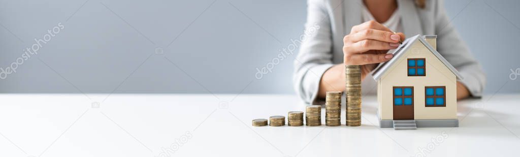 Person's Hand Inserting Coin In The House Model With Increasing Coins Stack On Desk