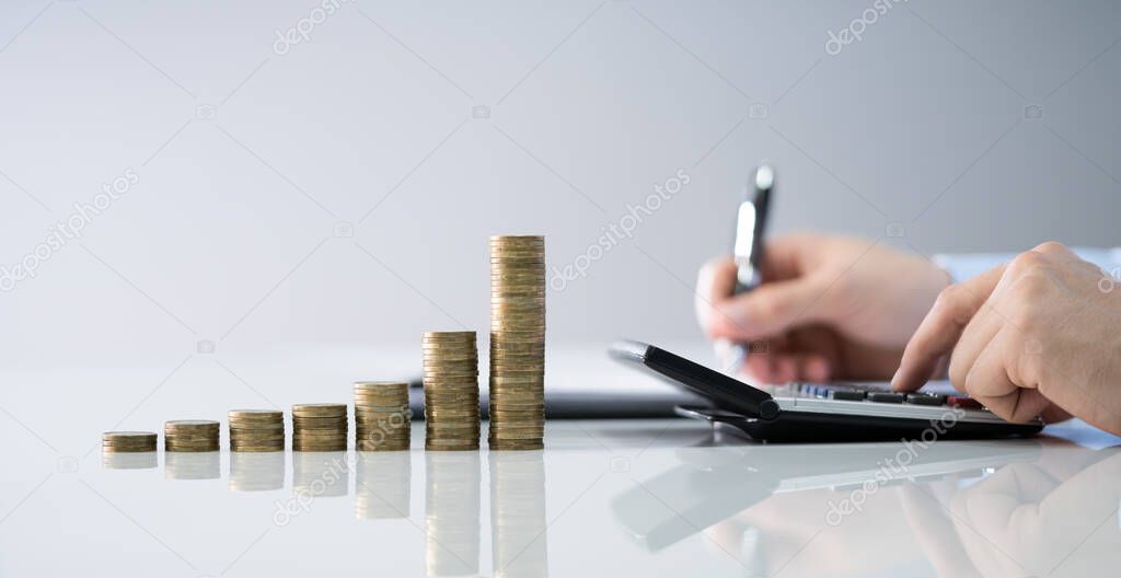 Businessman Calculating Invoice With Stacked Coins Arranged At Office Desk
