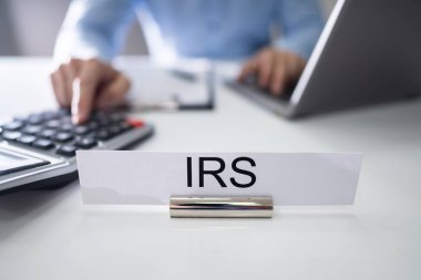 IRS tabela önünde muhasebeci hesap makinesi ve Laptop ile masa üzerinde vergi hesaplama