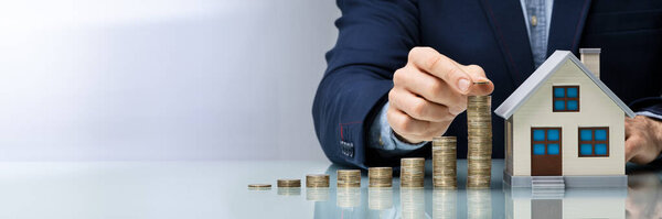 Close-up Of Man's Hand Stacking Coins Near House Model On Wooden Desk 