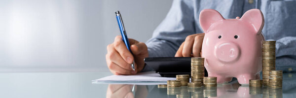 Close-up Of Businessman Calculating Bill With Piggybank And Stacked Coins On Desk In Office