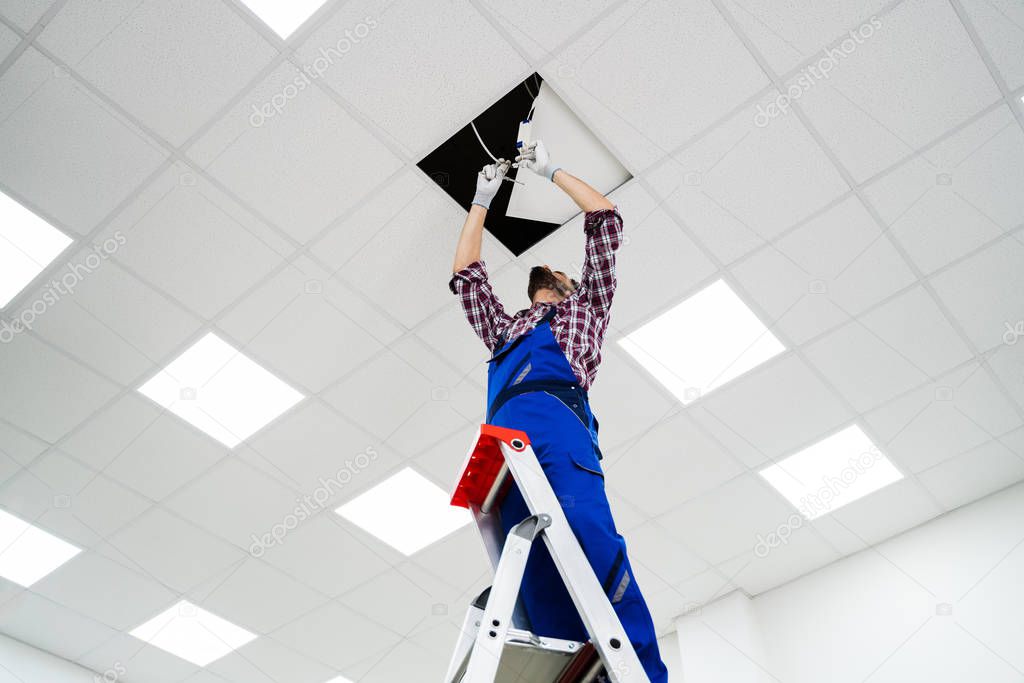 Full Length Portrait Of Electrician On Stepladder Installs Lighting To The Ceiling In Office