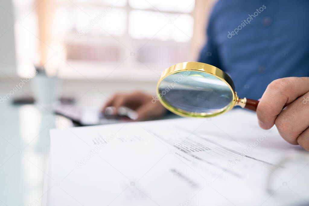 Close-up Of Businessperson Hand Examining Bills With Magnifying Glass At Desk